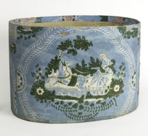 Image shows a bandbox bottom, with lid removed, covered in wallpaper with printed scene of a woman driving a chariot pulled by a single horse. Scene is enframed in a foliate scroll medallion. Please scroll down for a further description of this image.