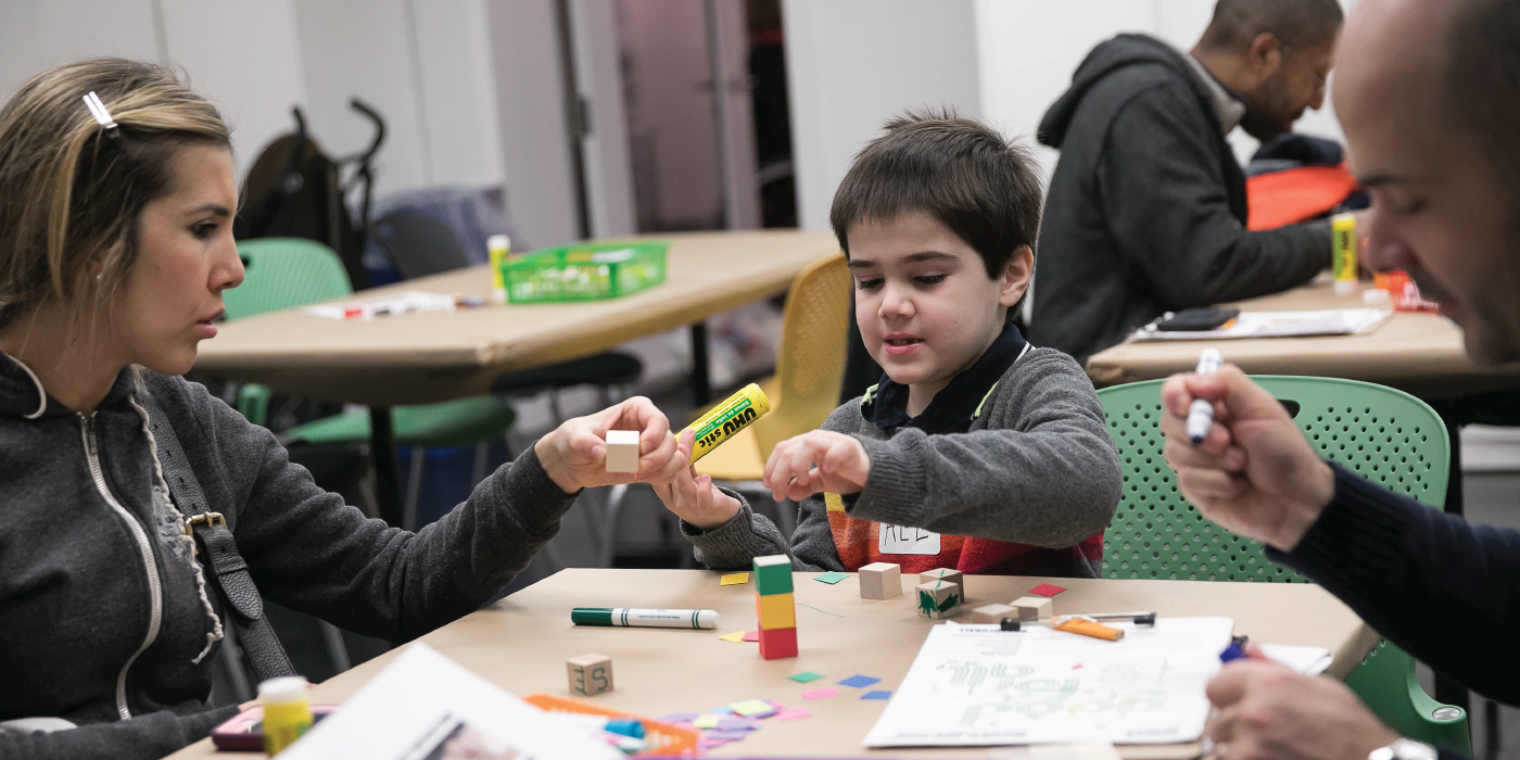 Young boy sits at table with colored blocks and parents seated on either side.