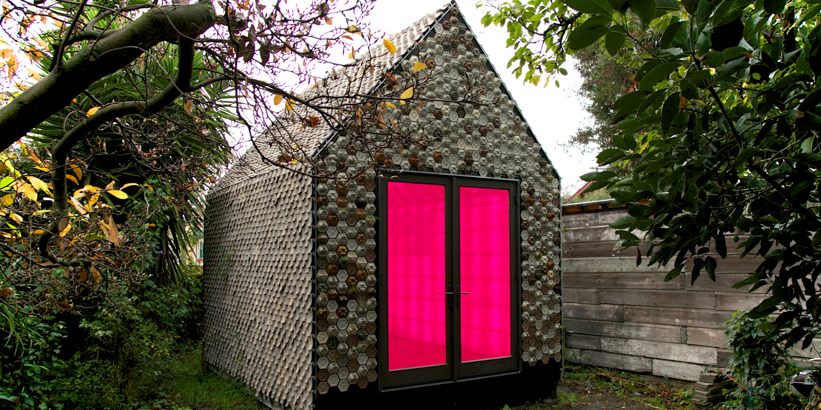 Diseno Lecture with Ronald Rael. Image of a modern cabin, constructed from small succulents. Bright pink doors illuminate on the front facade. Scroll down for additional program information.