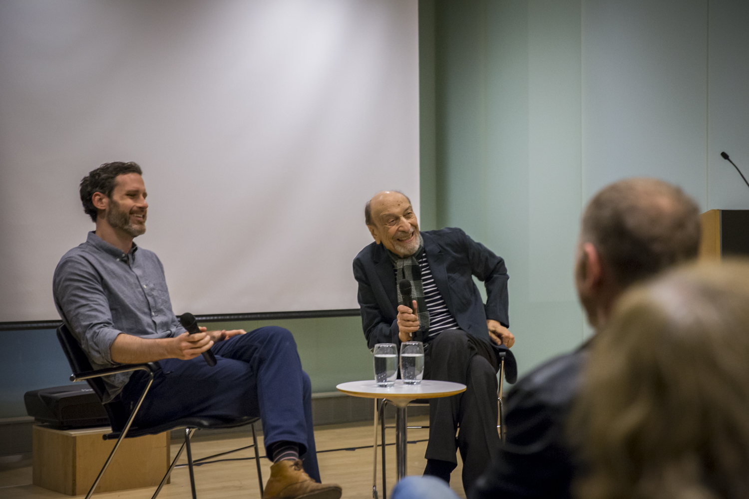 Image of two men on a stage, Milton Glaser is on the right, he holds a microphone and is laughing.