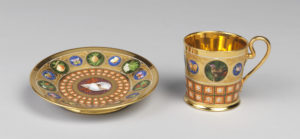 Verbal description tour of Tablescapes: Designs for Dining. Image of a gold mug and saucer made from ceramic with detailed colorful depictions of flowers, animals, and shells around the rim of each object.