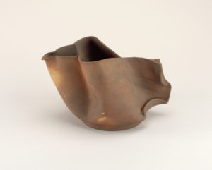 Image features short pitcher of unglazed clay in tones of tan to brown, the thin-walled body rippled and folded into an irregular form with short spout at one side, tab-like handle at opposite side. Please scroll down to read the blog post about this object.