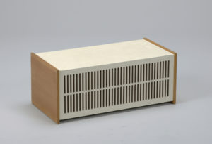 Image features an off-white rectangular speaker, the front with two rows of vertical slits; left and right sides faced with square, blond wood panels. Please scroll down to read the blog post about this object.