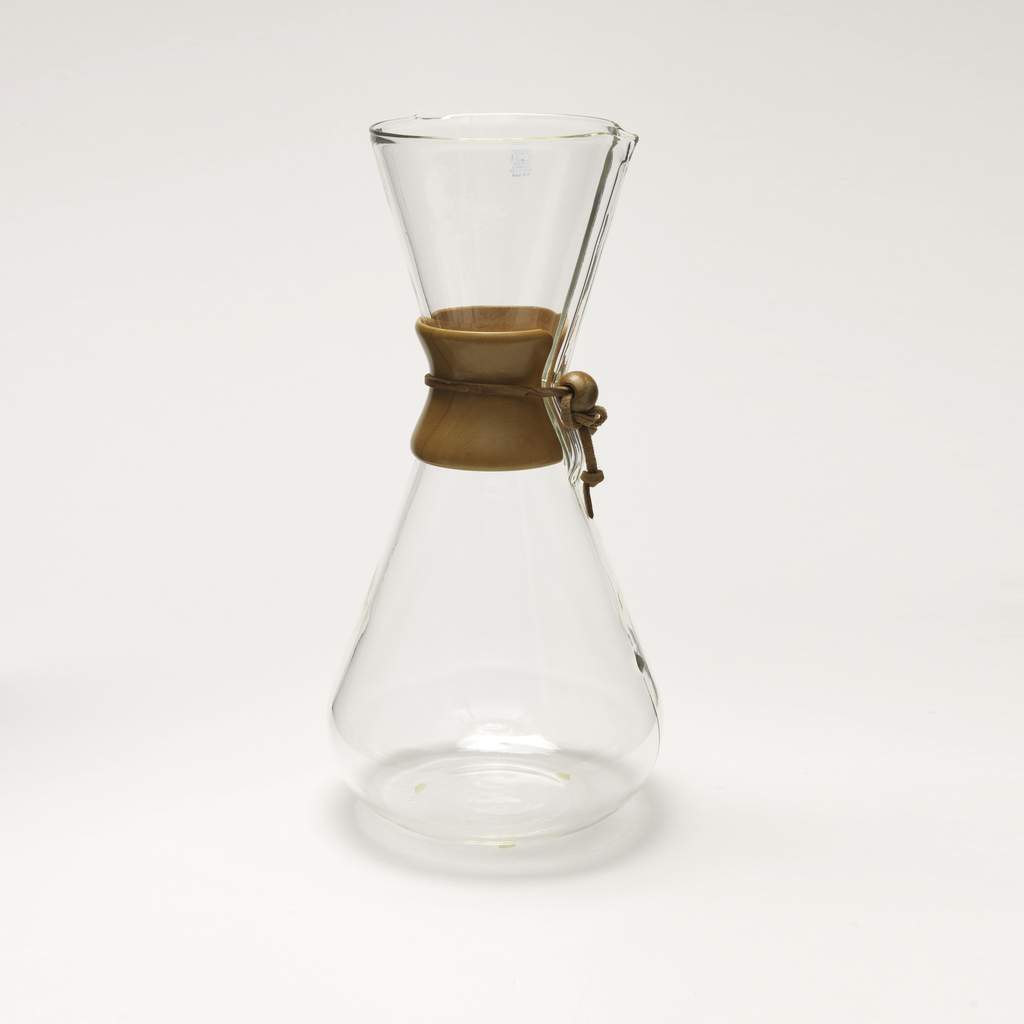 Image features an hourglass-shaped coffee maker of transparent glass with high neck and circular mouth molded with a narrow spout; tapered wood collar/hand grip at neck tied with leather thong with bead stop; small projecting dot as water level indicator in lower body, vertically aligned with spout. Please scroll down to read the blog post about this object.
