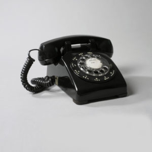 Image features telephone comprising wedge-shaped black plastic body, the front with clear circular rotary dial with finger holes and surrounded by white numerals and letters; handset with earpiece at one end and speaker at other, set in cradle at top rear of telephone body; coiled black plastic-covered cord connects handset to body. Please scroll down to read the blog post about this object.