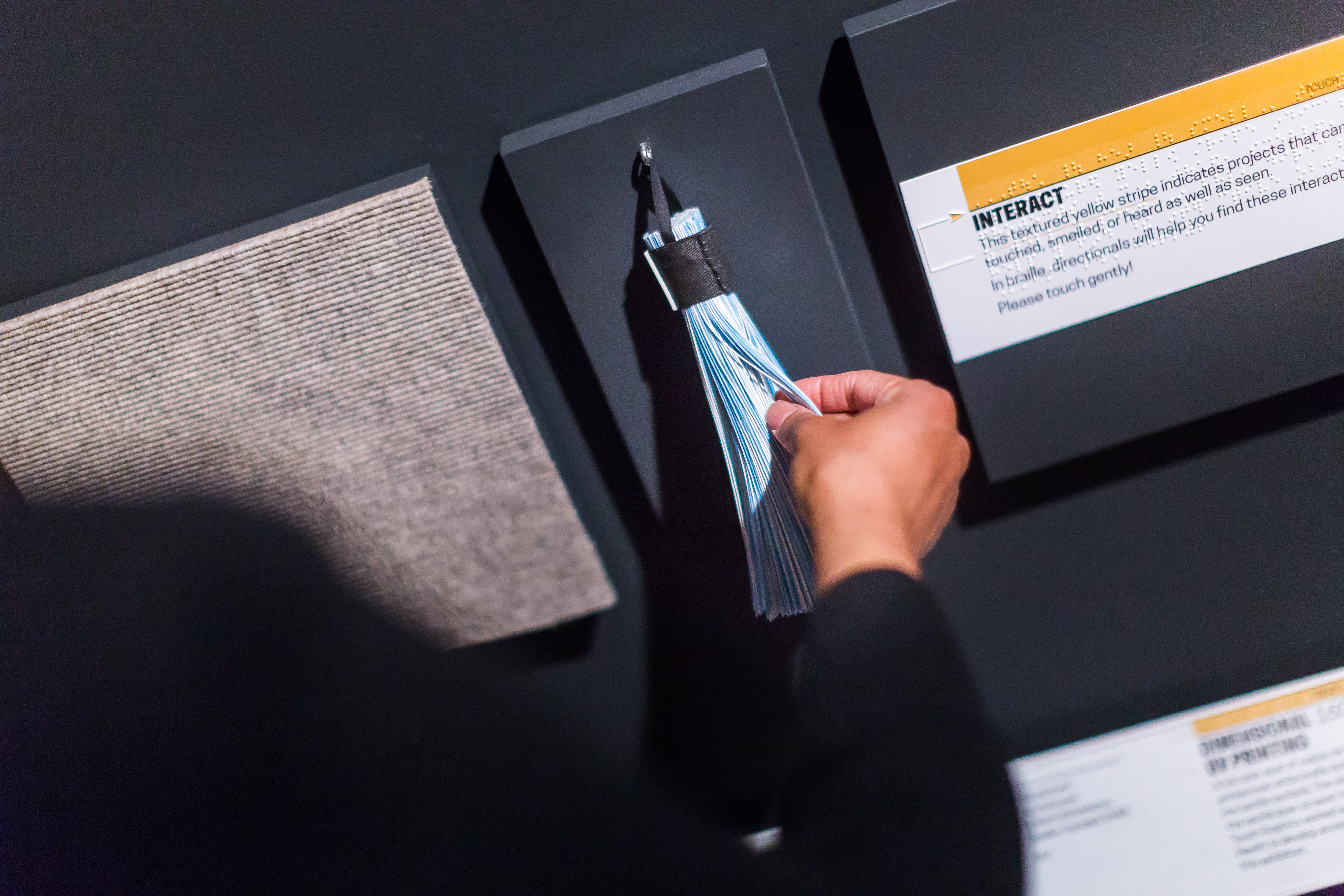 Close up photograph of visitor hand touching a small blue tassel adhered to the wall in Senses exhibition.