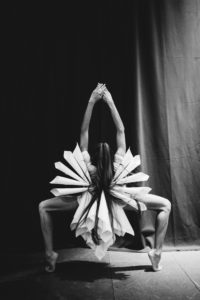 A black and white photograph of a dancer whose back is to us with her arms outstretched above her head, hands clasped, and her legs facing outwards, forming a rectangular shape as she stands on pointed toes. She wears an elaborate white costume made up of multiple cones pointing inwards, her long dark hair in a high ponytail falling well below her waist.