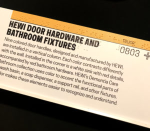 Horizontal photograph of an exhibition label on a rail. The labels are printed with a special process called dimensional UV printing. A full-color layer of visible ink is printed over white braille dots and raised-line tactile symbols. The visible layer and the tactile layer co-exist for all users.