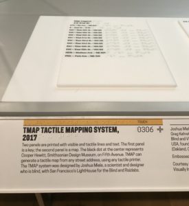 Vertical photograph of a museum gallery. On a table is a sheet that lists components of a map in both Latin text and braille. The accompanying rail states "TMAP Tactile Mapping System, 2017" with additional descriptive text about the other objects on the table. The label combines both Latin text and braille.