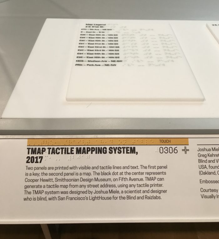 Vertical photograph of a museum gallery. On a table is a sheet that lists components of a map in both Latin text and braille. The accompanying rail states 