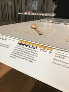 Vertical photograph of a museum gallery. A spoon with bumpy edges sits upon a table. The accompanying rail states "Dining Toys, 2017" with additional descriptive text about the other objects on the table. The label combines both Latin text and braille.