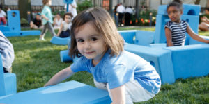 A girl in the foreground holds a big blue block with both hands, while a girl in the background sits amidst several large blue blocks.