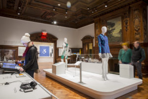 Photograph of the exhibition Access+Ability. Several visitors view objects displayed on mannequins in the center of a large room. One interacts with a touchscreen tablet, the international symbol of accessibility is visible on the far back wall. Scroll down for information about a verbal description and sensory tour of the exhibition.