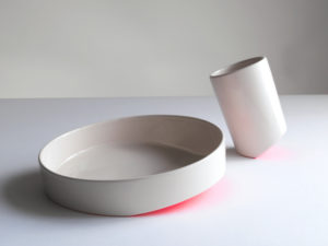 A white plate and cup illuminated from the bottom by a pinkish-red light from the Share.Food Tableware collection, 2013; Bilge Nur Saltik (Turkish, b. 1988), Studio Bilge Nur Saltik (London, UK, founded 2013); Ceramic; © Studio Bilge Nur Saltik. On view in The Senses: Design Beyond Vision.