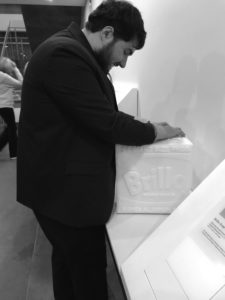 Shown here is a photograph of Sina at the Andy Warhol Museum in Pittsburgh. Sina is a young man with dark hair and a trim beard. He is touching a tactile model of one of Andy Warhol’s famous Brillo Box sculptures. The model is white. It has a raised surface that reproduces the lettering and graphics on Warhol’s sculpture.