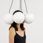 A woman standing with her head surrounded by giant white orbs dangling from black cords. Tactile Headset, 2014–18; Alessandro Perini. On view in The Senses: Design Beyond Vision.