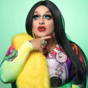 Portrait photo of light/medium skin-toned drag queen Horrorchata in an intentionally coquettish pose, with long, black, straight hair and artificially fluorescent blue eyes. She wears a brightly colored, skin-tight, airbrush-style shirt, with a canary yellow faux fur stole over her right shoulder. She has dramatically made-up eyes and cherry-red lips, evocative of chola contemporary style, and is adorned with oversized jewelry. Her portrait is set against a glowing aqua-colored background.