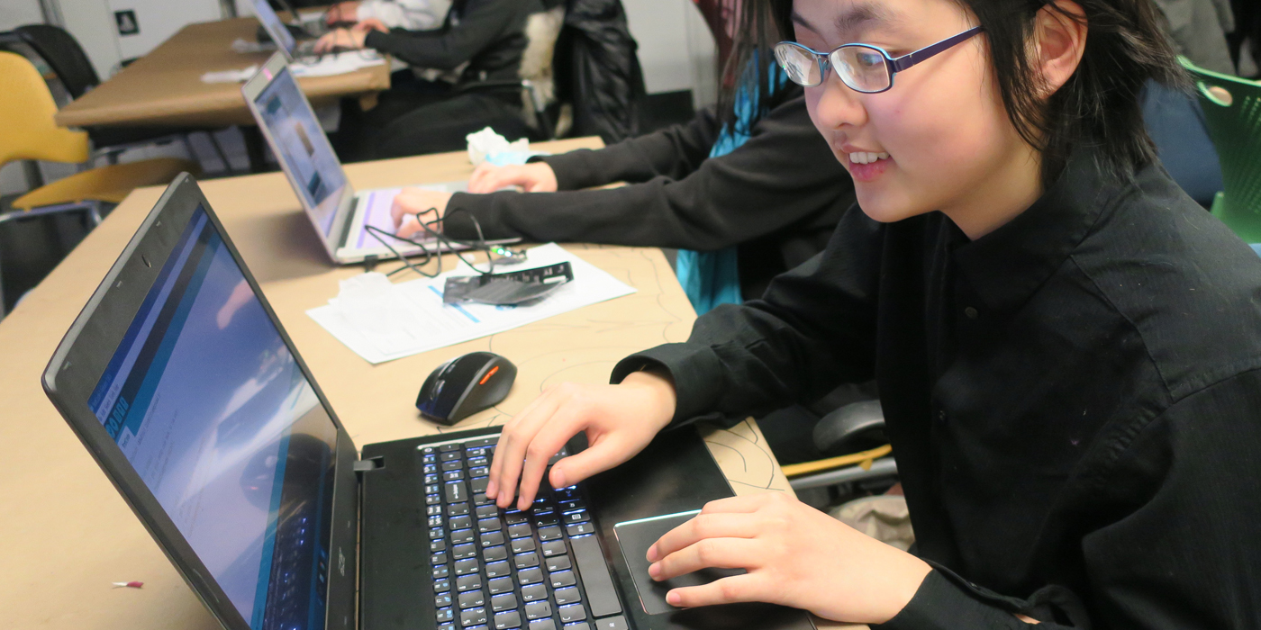 A girl wearing glasses smiles while looking at the laptop computer screen in front of her. Her hands are on the keyboard and track pad.
