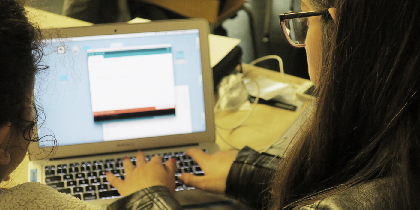 Two girls are seen from the back as they type on a coding program on a laptop computer.