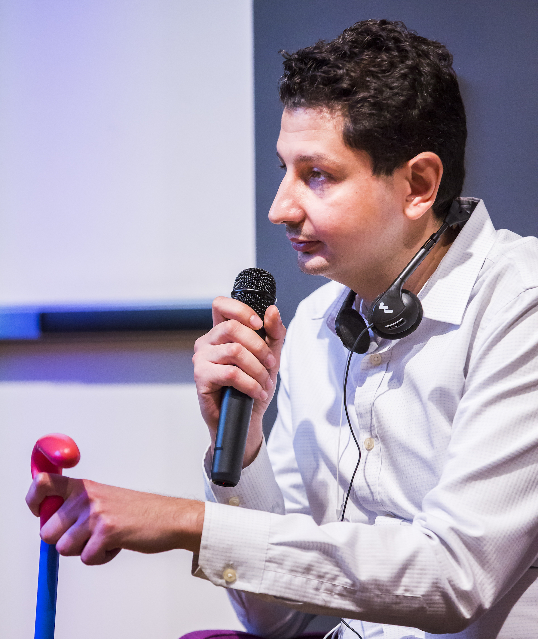 Photo of Walei Subray. He is seated, and holds a microphone in one hand and a cane in the other. He has a thoughtful expression.