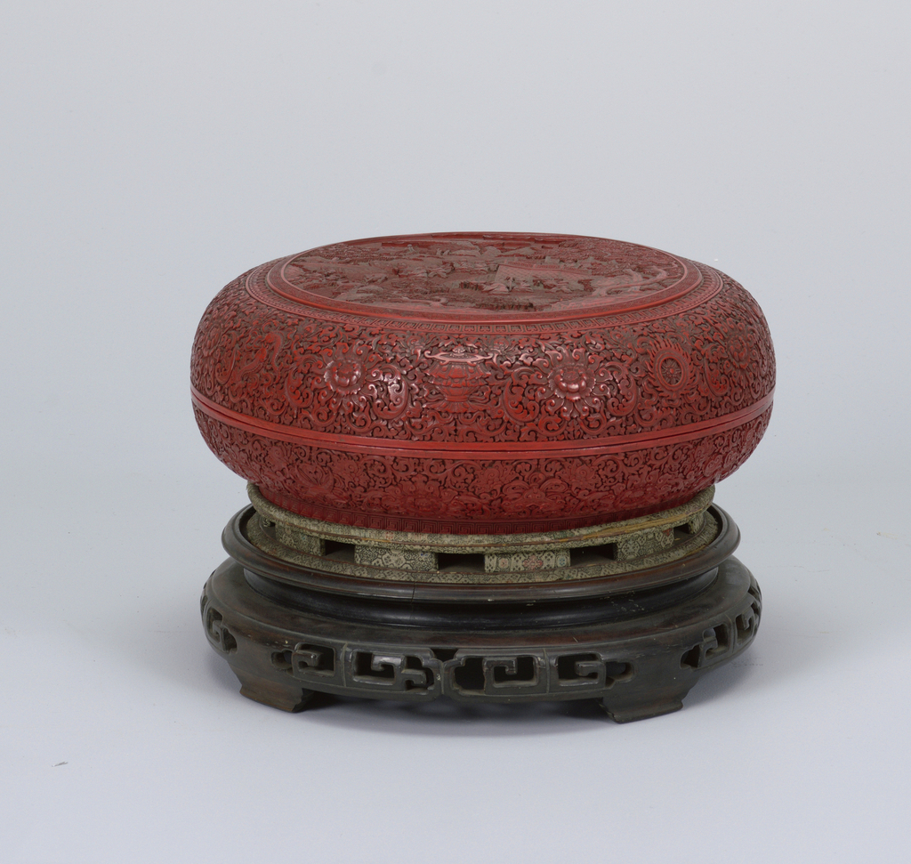 Image features a low, deep red circular box deeply carved with a landscape on the lid, surrounded by bands and fields of geometric and floral decoration and Buddhist symbols. Box sits on a carved circular wooden stand. Please scroll down to read the blog post about this object.