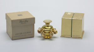 Image features squat round clear glass bottle encased in a gold-toned metal sleeve with six jingling bells around circumference; gold-toned flaring circular foot; removable circular gold-toned metal cap. Separate square, gold foil-covered presentation box and brown cardboard outer box. Please scroll down to read the blog post about this object.