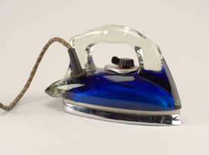 Image features iron with clear glass housing encasing blue emulsion-covered body, with glass handle and metal sole plate; plastic and metal control knob below handle; power cord at rear. Please scroll down read more about this object.