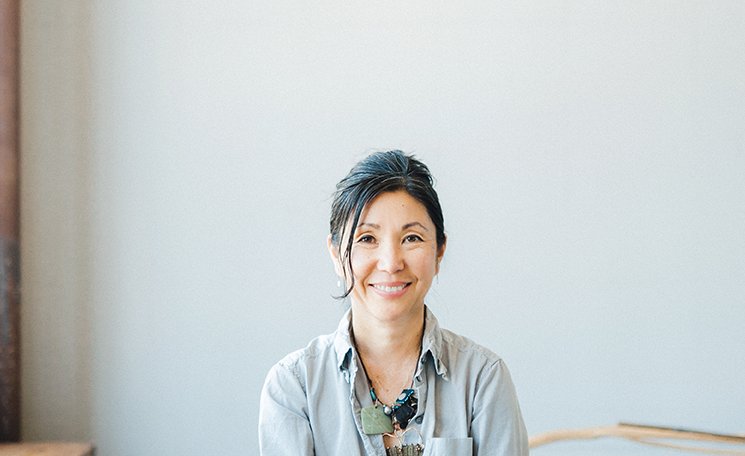A photograph of designer Christina Kim smiling at the camera. She wears a button-up shirt with a necklace made with large green and black stones.