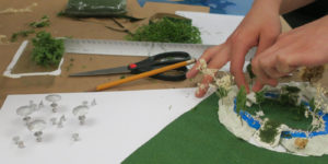 Two hands are seen at the side of the zoomed-in image. The hands are placing small plastic modeling trees onto an architectural model. In the left-hand side of the image, tiny silver modeling umbrellas are also visible.