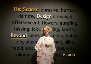 Ellen Lupton stands in front of the title wall for the exhibition The Senses: Design Beyond Vision