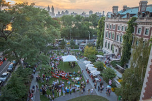 Aerial view of south-facing facade of Carnegie Mansion and Cooper Hewitt's Arthur Ross Terrace and Garden at sunset in summer. A crowd has gathered on the lawn around a white tent. Inside the tent musicians are performing. Central Park and the Jacqueline Kennedy Onassis Reservoir are visible in the distance.