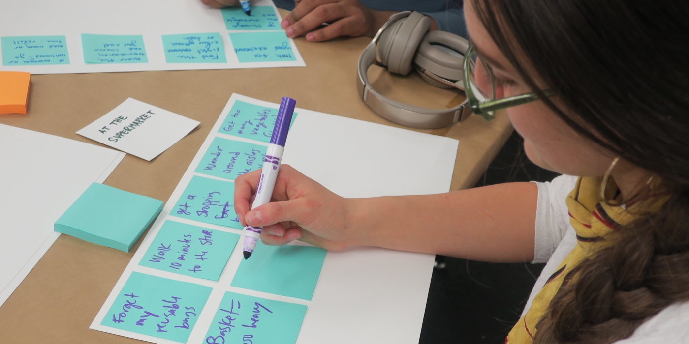 A young woman wearing glasses writes on a post-it note, itself attached to a larger piece of paper.