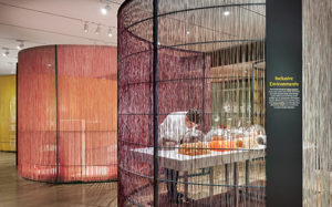 A museum gallery is filled with large, undulating partitions composed of colorful threads. Through the nearest partition, a man can be seen leaning forward to smell scented objects housed in glass domes. A sign on this partition reads "Inclusive Environments".