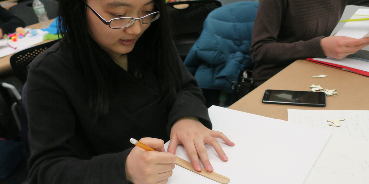 A girl looks down in concentration. She is tracing the outline of a wooden tongue depressor on a white sheet of paper.