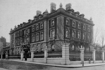 The image is a photograph from 1903 of the Carnegie Mansion. It is black and white. The building is photographed from the street from the north west. A fence with large posts surrounds and rectangular, block-like building. It is four floors with many windows. Decorative urns and three chimneys are stationed arond the roof. The three tress along the right side of the photograph do not have leaves.