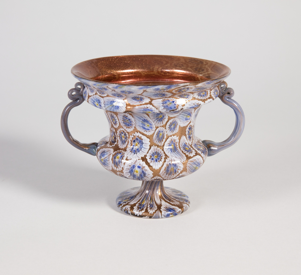 Image features short glass vase of of campana-urn form, the interior of copper-toned aventurine the exterior with blue, white and yellow florette murrines; C-form handles on left and right. Please scroll down to read the blog post about this object.