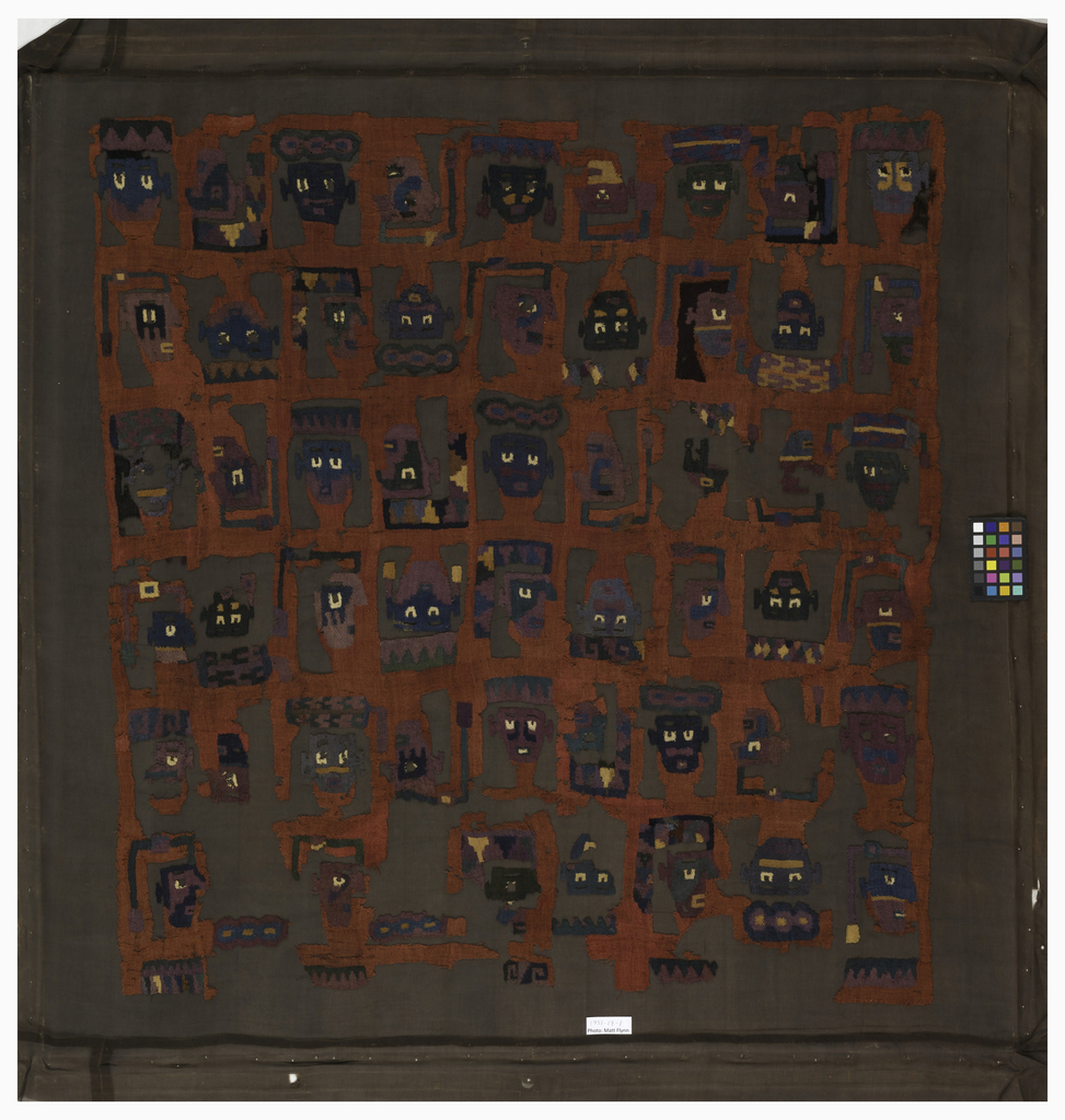 Image features: Fragment of a mantle with close-set horizontal rows of stylized warriors' heads, each with different headdress and color combinations. In the top row, upright front-facing heads alternate with upside down heads in profile. In the next row, upright heads in profile alternate with upside down front-facing heads. Rich muted shades of brown, gold, tan, blue, green, purple, and white on a rust-red ground. Please scroll down to read the blog post about this object.