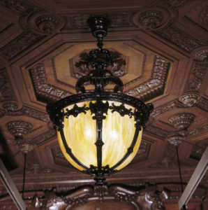 The image is a photograph of a light fixture hanging from a ceiling. The ceiling is ornately carved wood divided into octagons with foliate details. The light fixture itself extends down about three feet from the ceiling. The structure is composed of a dark polished metal. The luminous portion (about half of the oval structure) is yellow and bulbous.