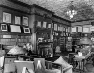 The image is a photograph picturing an office as it looked in 1911. The photograph is black and white. Chairs and sofas are nestled amongst tables, the latter of which are covered in textiles and various framed documents. A fireplace is in the center of the image. Bookshelves line the bottom half of the walls while more framed documents line the top half. A small portion of the walls below the ceiling contain a painted panel with a scroll and inspirational quotes.