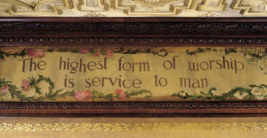 The image is a photograph of a painted panel positioned along a ceiling. It is framed in carved wood molding. The panel depicts a trompe l'oeil scroll containing the words "The hightest form of worship is service to man." The scroll is bordered with leafy vines and pink and white flowers.