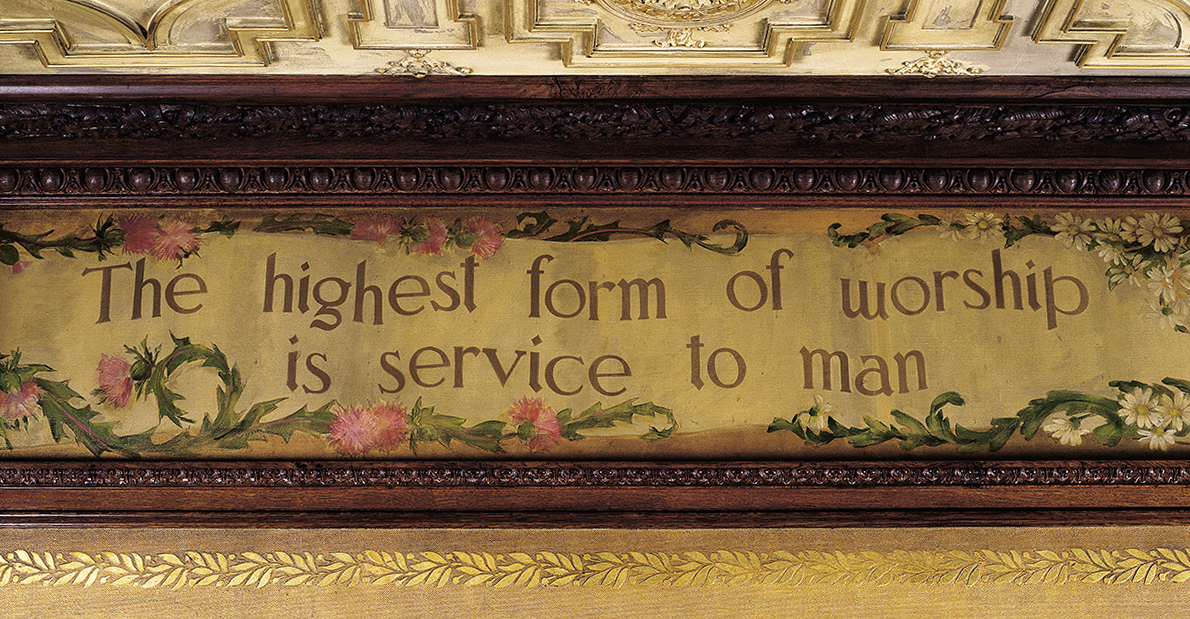 The image is a photograph of a painted panel positioned along a ceiling. It is framed in carved wood molding. The panel depicts a trompe l'oeil scroll containing the words 