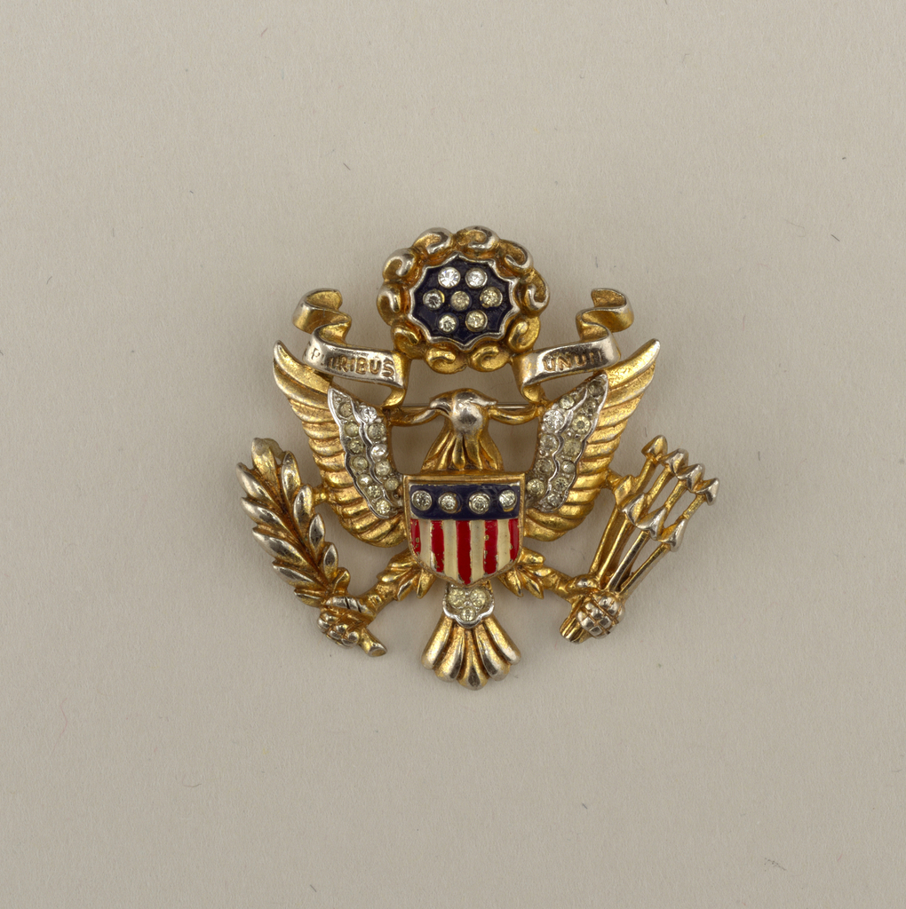 Image features gold toned metal brooch with red white and blue enamel and glass paste decoration depicting the great seal of the United States: a shield supported by a bald eagle, wings and legs outstretched, holding a scroll in its beak with the motto “E PLURIBUS UNUM,” and grasping an olive branch in its right talon and a bundle of arrows in its left talon; above its head a scalloped blue circle with 6 glass pastes and gold toned surround. Please scroll down to read the blog post about this object.