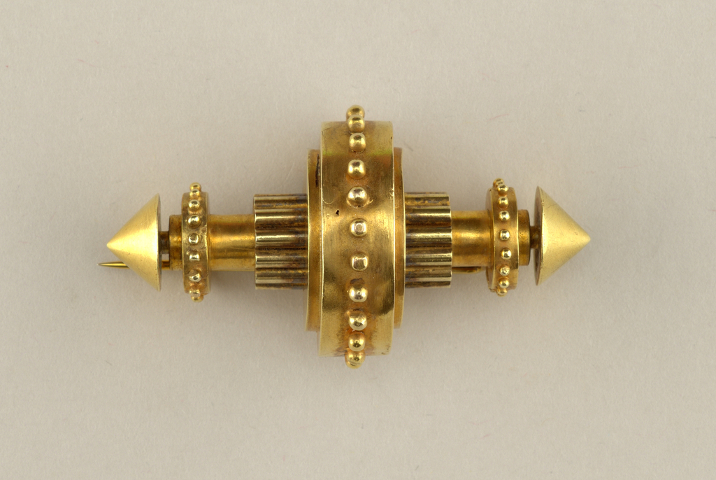 Image features a gold brooch of symmetrical geometric form reminiscent of a machine part; composed of a central shaft with two sets of ten small cylinders bundled around the center, encircled by a large beaded band at the center; conical terminals situated at each end of the central shaft with small beaded bands just inside. Please scroll down to read the blog post about this object.