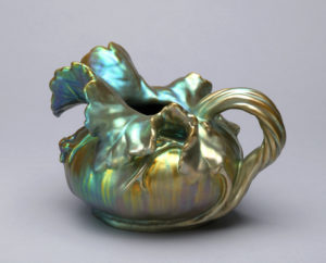 A pitcher of squat bulbous gourd form with a large leaf-shaped spout and handle in the form of a gathered root or vine; green and gold iridescent luster finish.