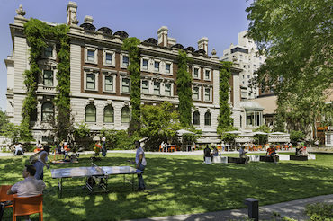 The facade of the Carnegie Mansion with wisteria climbing up the facade and fronted by a spacious green lawn where people are playing ping-pong and relaxing.