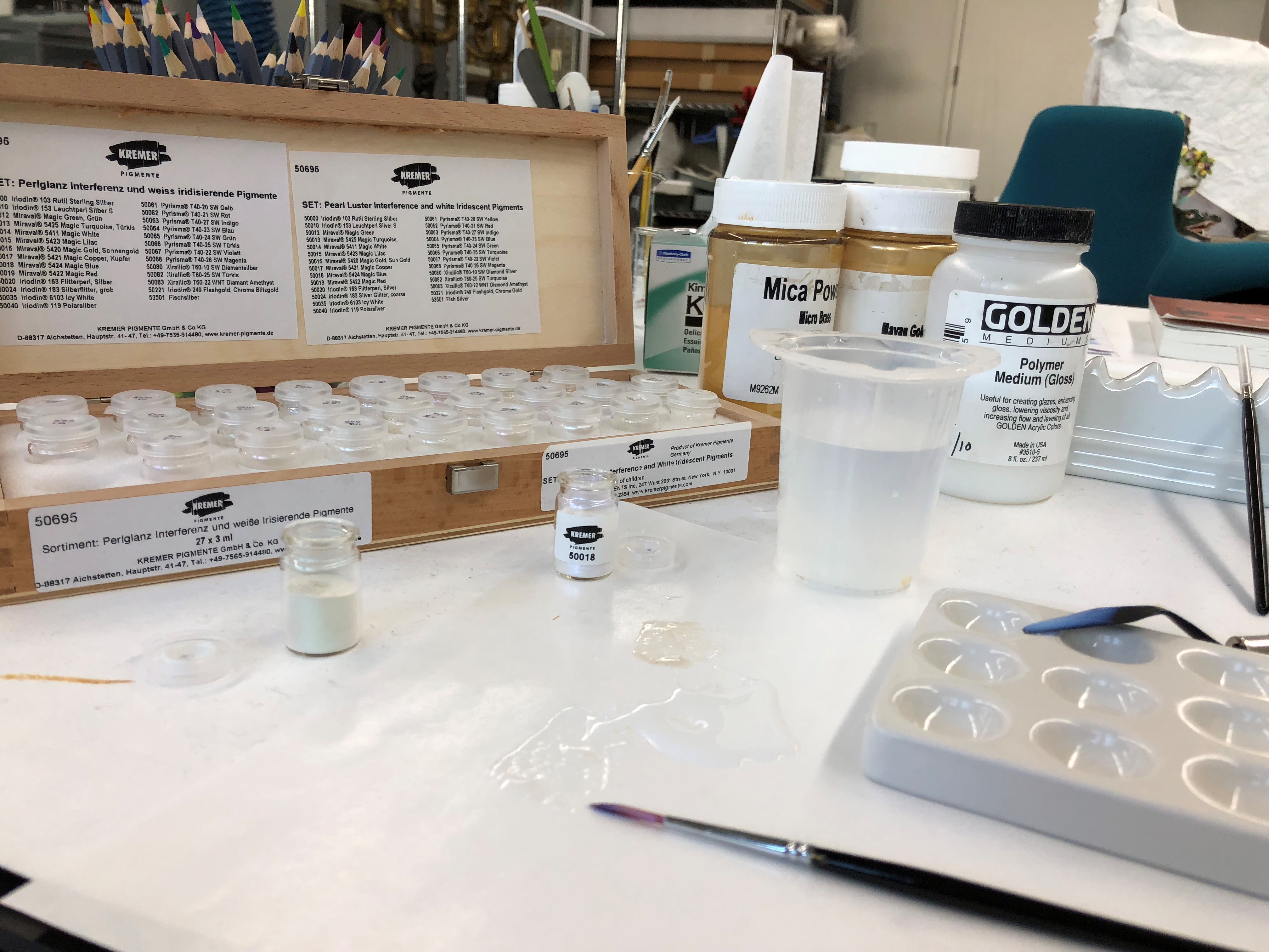 Photograph of a tabletop in a lab. It pictures a box of small viles containing pigrments. Various cups, jars, and brushes are placed around the workspace.