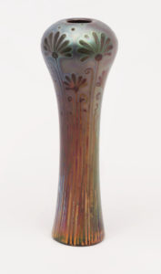 Image features tall, slender, slightly cylindrical vase with globular shoulder, no neck, incurving rim; no foot. Design of tall flowers (stylized carnations) and curving leaves that rise up onto the shoulder. Beneath and between these are smaller flowers and leaves. A few random dots on top of shoulder. Decoration in metallic lustres on an iridescent ground, in shades of peacock blue, lavender, crimson, and green against a copper glaze ground. Please scroll down to read the blog post about this object.