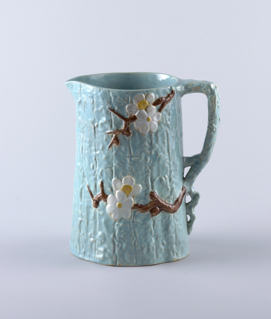 Image features a conical pitcher, molded to resemble a tree trunk, the handle in the form of a gnarled branch. Body glazed with a light blue ground decorated with brown branches bearing white and yellow blossoms. Please scroll down to read the blog post about this object.