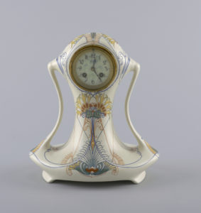 Image features table clock in curving glazed earthenware case with polychrome linear decoration of stylized plant forms on a cream-colored ground. Please scroll down to read the blog post about this object.