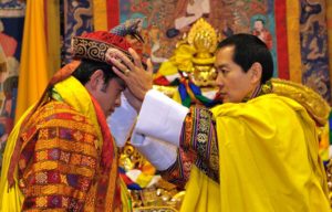 A man in a brilliant yellow robe places a crown on the head of a man in a multi-colored patterned robe.